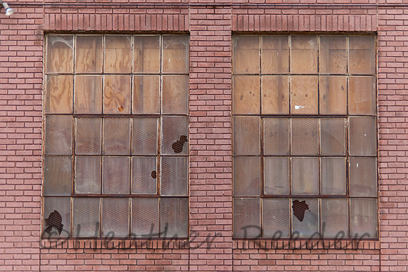Two boarded and broken windows on aged red brick wall.